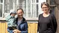 Tre personer, varav en vuxen håller ett litet barn, står framför en gul trävägg med ett fönster med spetsgardiner.