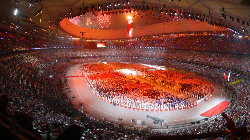 Komeasti punaisella ja valkoisella valaistu olympiastadion sisältä, keskellä nurmella iso joukko esiintyjiä.