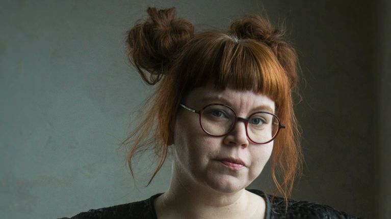 En mörkklädd kvinna (konstnären Alexandra Furubacka) med rött uppsatt hår framför neutral bakgrund.
