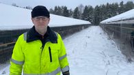 En man med glasögon och mössa iklädd reflexjacka står i snön mellan två rävhus på en pälsfarm. 
