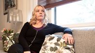 Nainen istuu harmaalla sohvalla kukkakuvioisten tyynyjen välissä olohuoneessa, taustalla ikkuna ja lamppu.