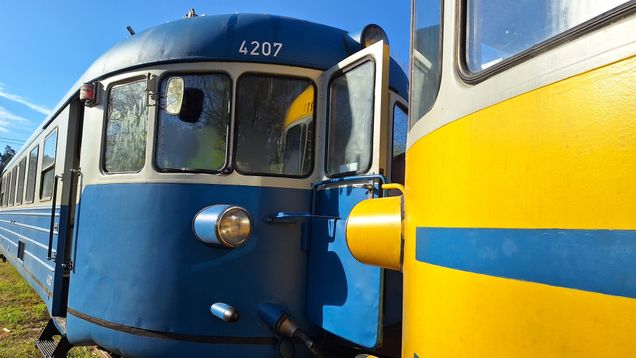 Två blågrå rälsbussar vid en bangård i soligt väder.