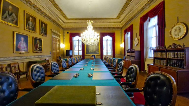 Det här är ett elegant mötesrum med en lång konferensbord dekorerat med ljuskrona. Väggarna pryds av porträtt och det finns hyllor med böcker och andra dekorativa föremål.