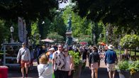Many tourists walking and posing for photos on a sunny summer day in a park with a greenish statue in the background.