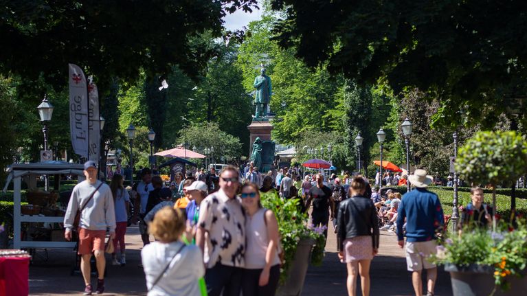 Many tourists walking and posing for photos on a sunny summer day in a park with a greenish statue in the background.