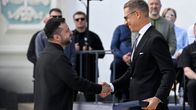 Alexander Stubb ja Volodymyr Zelenskyi kättelee ulkoilmatilaisuudessa, Stubb pitää kädessään laatikkoa. Taustalla seisoo ja istuu useita ihmisiä.