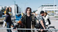 En person i mönstrad rock står vid ett staket på en gata med byggarbete, medan fotgängare och cyklister passerar bakom honom.