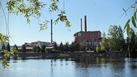 Punaruskea vaneritehdas piippuineen. Tehtaan etualalla väreilee Pielisjoki. 