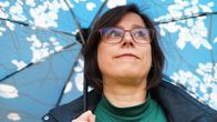 Tummahiuksinen ja sinitakkinen nainen katsoo yläviistoon sinivalkea sateenvarjo kädessäään. 
