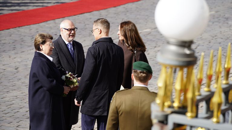 Two women and two men stand conferring on a cobblestoned square with gilt-topped fence and a uniformed officer in the foreground.