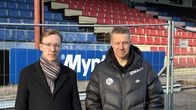 Två män i mörka överrockar står framför en fotbollsläktare i trä.