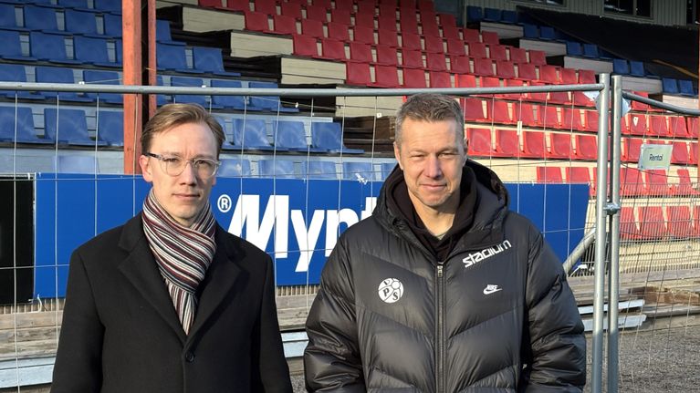 Två män i mörka överrockar står framför en fotbollsläktare i trä.