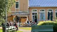 Ihmisiä istuu terassilla vaalean puurakennuksen edessä aurinkoisessa Viikinsaaren maisemassa.