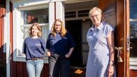 Två unga kvinnor klädda i jeans och likadana t-skjortor, samt en äldre kvinna i blåvitrandig klänning. De står på trappan framför ett rött trähus.