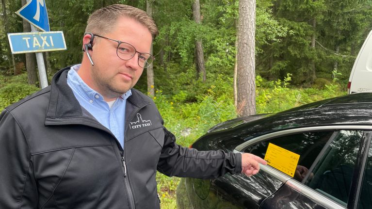En man med glasögon och headset pekar på en gul lapp på en svart bil.