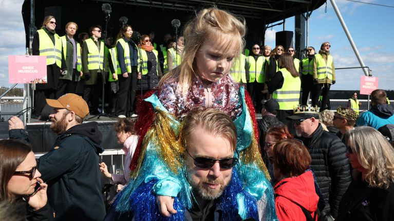 Lapsi istuu aikuisen hartioilla, taustalla laulaa huomioliiveihin pukeutunut kuoro.