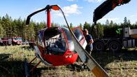 Punainen helikopteri on maassa metsäaukealla, siipi vaurioitunut. 