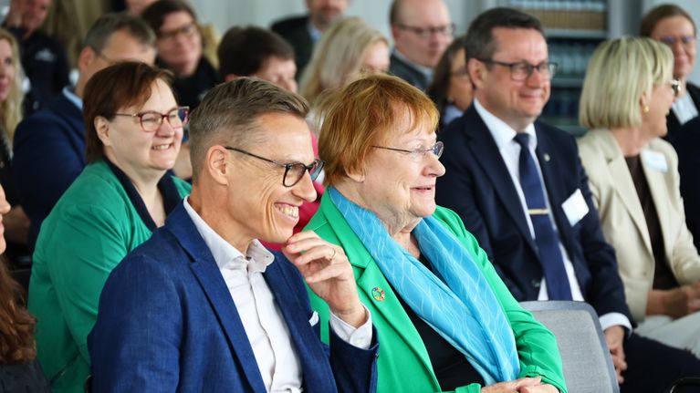 Middle-aged people, including Alexander Stubb and Tarja Halonen, sitting next to each other at a conference. 