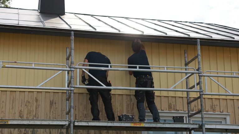 Två personer arbetar på en byggnadsställning framför ett gult trähus.