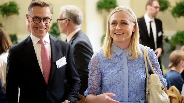 A tall man in a black suit and pink tie stands next to a blonde woman in a blue blouse, both smiling.