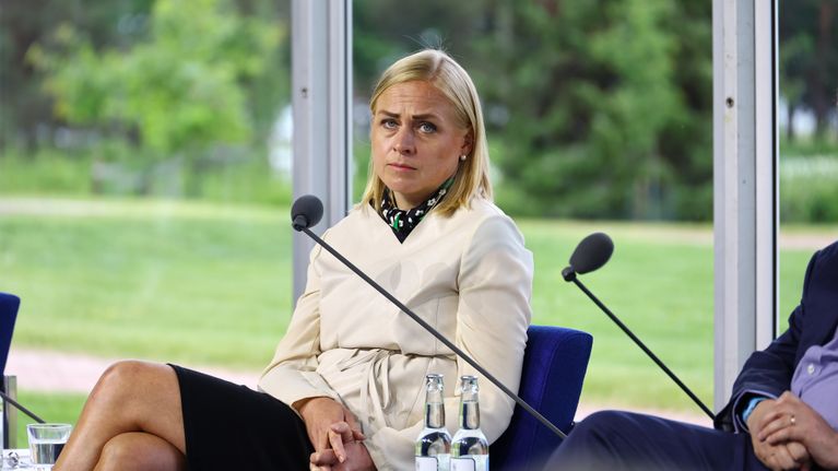 Woman with blonde hair wearing beige jacket, sitting on a chair behind a microphone, with trees and grass in the background.