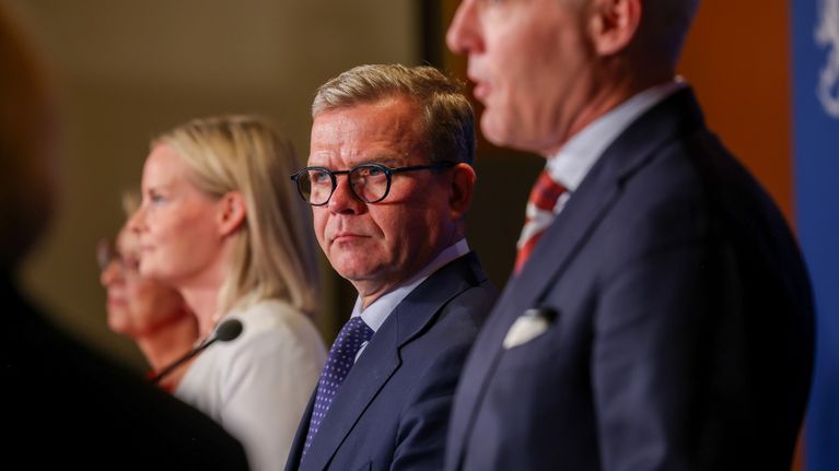Neljä ministeria seisoo rivissä mikrofonien edessä sisätilassa.