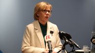 Anna-Maja Henriksson, with blonde hair and white jacket over a green blouse, speaking into microphones at a podium.