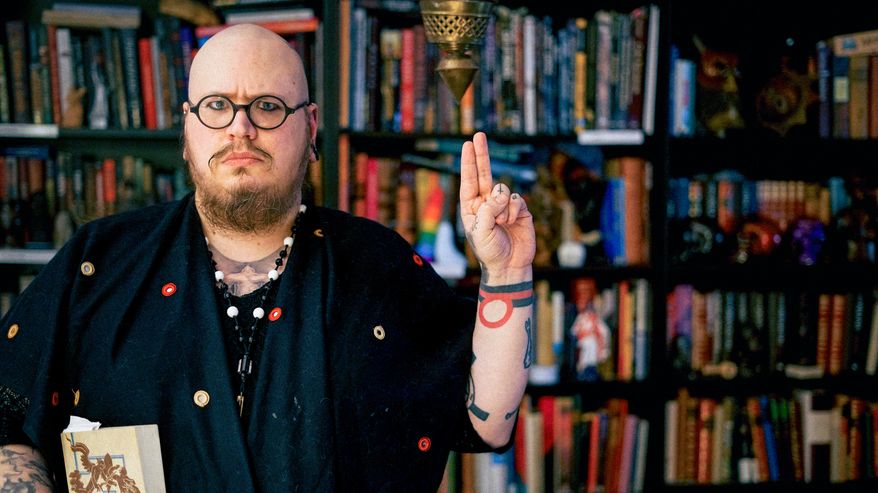 Henri Hiljander katsoo vakavana kameraan ja näyttää käsimerkkiä, jossa etu- ja keskisormi ovat pystyssä vierekkäin.