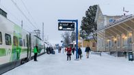 Tågresenärer står vid perrongen bredvid ett tåg som väntar att avgå. Det är snö på marken och stationsbyggnaden syns till höger. 