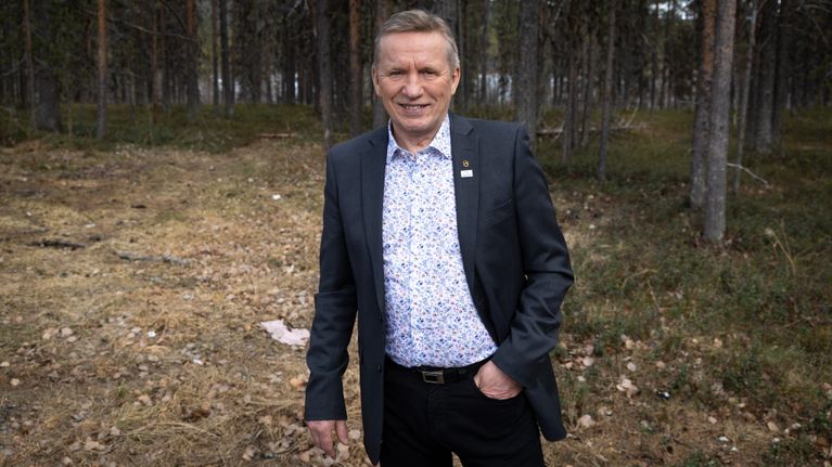 Tummapukuinen mies hymyilee metsän reunalla.
