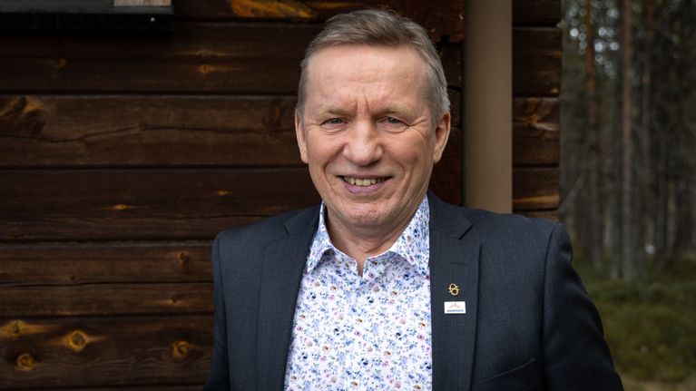 Tummapukuinen mies hymyilee metsän reunalla, tumma hirsirakennus taustallaan.