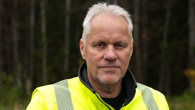 Työtakkinen mies seisoo metsän reunalla ja katsoo kameraan.