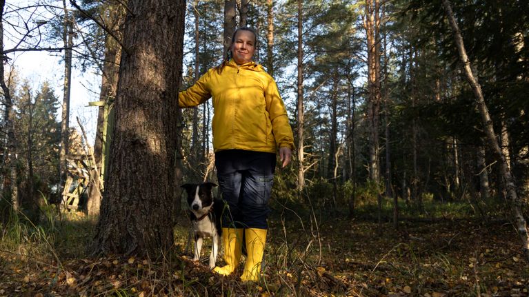 Hymyilevä nainen keltaisessa takissa ja kumisaappaissa seisoo metsässä koiransa kanssa.