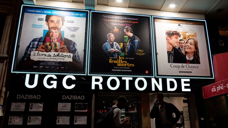 UGC Rotonde elokuvateatterin ovilla kaksi ihmistä puhelimillaan. Yläpuolellä kolmen elokuvan mainokset, joista keskimmäinen Aki Kaurismäen Kuolleet Lehdet ranskan kielisellä nimellä les feuilles mortes.