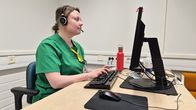 Sairaanhoitaja Mari Lappalainen kirjoittaa näppäimistöllä tietokoneen ääressä. Hänellä on  mikrofonilliset kuulokkeet päässään.