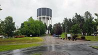 Lappeenrannan vesitorni kohoaa puiden takaa, etualalla soratie ja pysäköityjä autoja sekä opastekyltti.