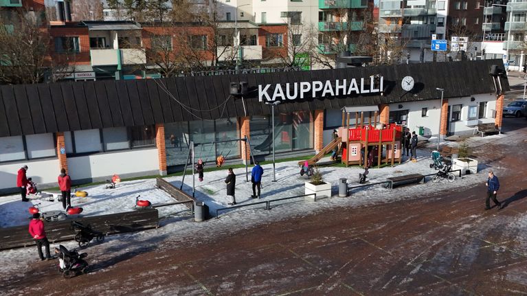 Ilmakuvassa on talvinen näkymä torialueella sijaitsevasta Lappeenrannan Kauppahallista, jonka edustalla on puistoalue, jossa lapset leikkivät lumessa.