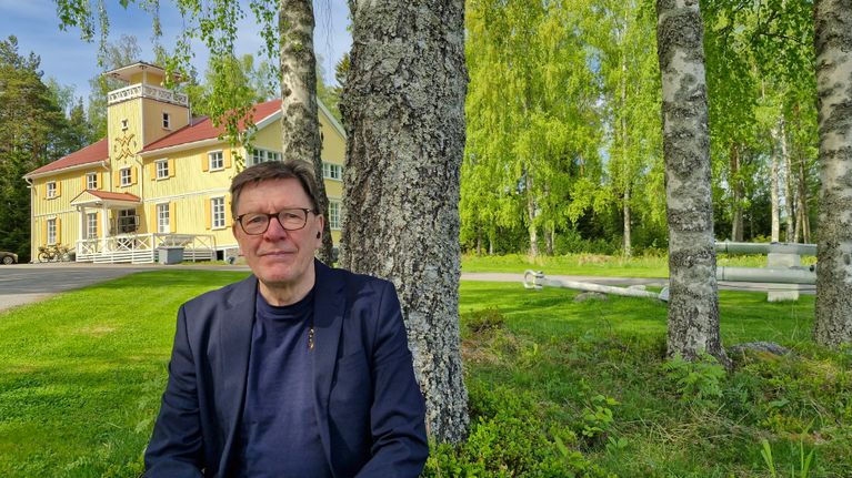 Silmälasipäinen mies pikkutakissa istumassa koivun juurella taustallaan vaasalaisen vanhan pursiseuran paviljonki. 