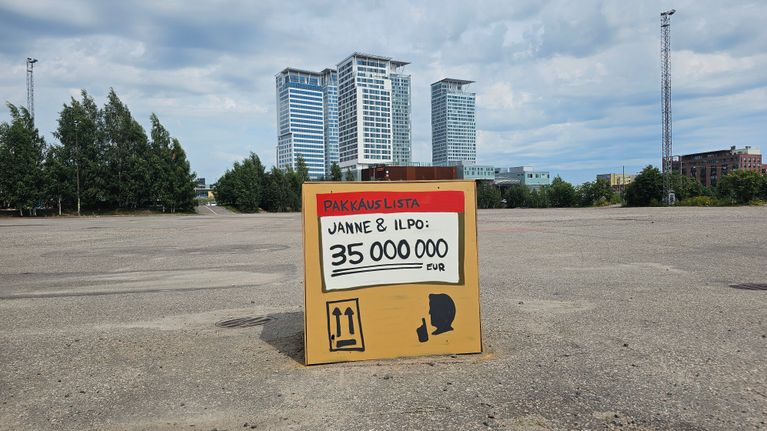 Tyhjällä asvalttikentällä on isokokoinen ruskea laatikko, jossa lukee “Pakkauslista, Janne & Ilpo: 35 000 000 EUR”. Taustalla korkeita rakennuksia.