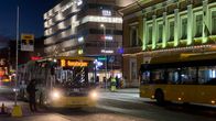 Tummiin vaatteisiin pukeutunut mies nousee bussiin. Bussin valot ovat kuvaajan suuntaan. Tien toisella puolella ohi ajaa toinen bussi poispäin kuvasta. Taustalla on KOP-kolmion rakennus jonka edessä on vihreisiin huomiovaatteisiin pukeutunut liikenteenohjaaja.