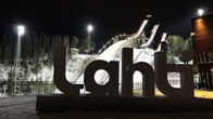 Lähikuvassa on tekstiteos, jossa lukee Lahti. Taustalla on lunta ja kolme korkeaa valaistua betonitornia, jotka ovat hyppyrimäkiä.
