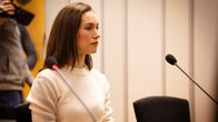 Woman with long dark hair, wearing a white sweater sitting at a desk with two microphones in a courtroom.
