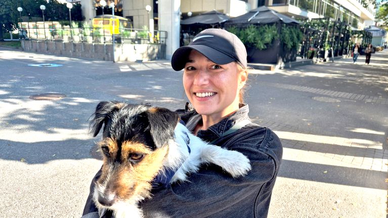En person med mörk keps håller en hund framför en större byggnad med uteservering och grönska.
