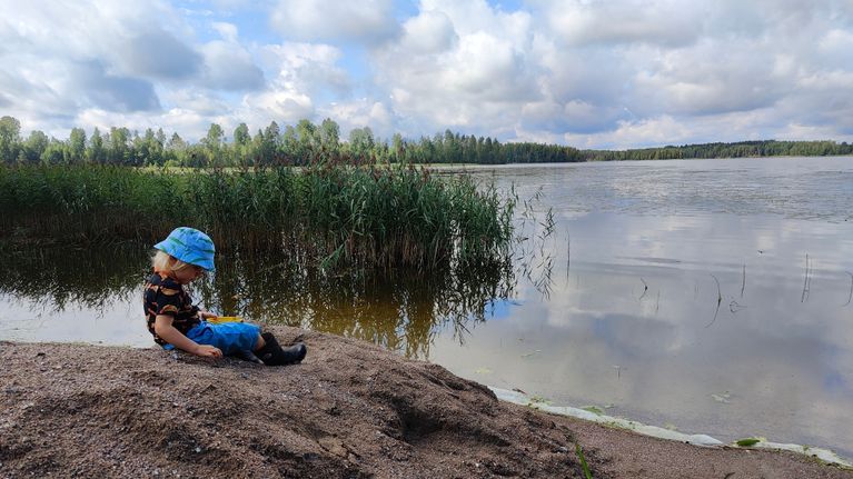 Pieni lapsi istuu hiekkakasan päällä järven rannalla. 