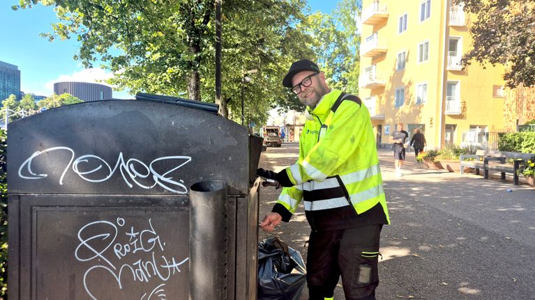 En person i varselkläder slänger skräp i en papperskorg på en stadsgata; texten "Resig Nadt" syns på papperskorgen.
