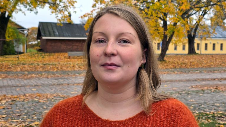 LAB:in asiantuntija Tiia Puolakka seisoo oranssi neule päällä ulkona, taustalla syksyisiä puita ja keltaisia rakennuksia.