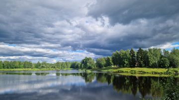Järvimaisema, jonka rantaa reunustavat vihreät puut ja taivaalla on tummia pilviä.