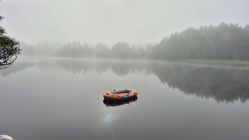 Oranssi ja musta kumivene kelluu sumuisella järvellä, taustalla näkyy metsää ja himmeästi aurinko.