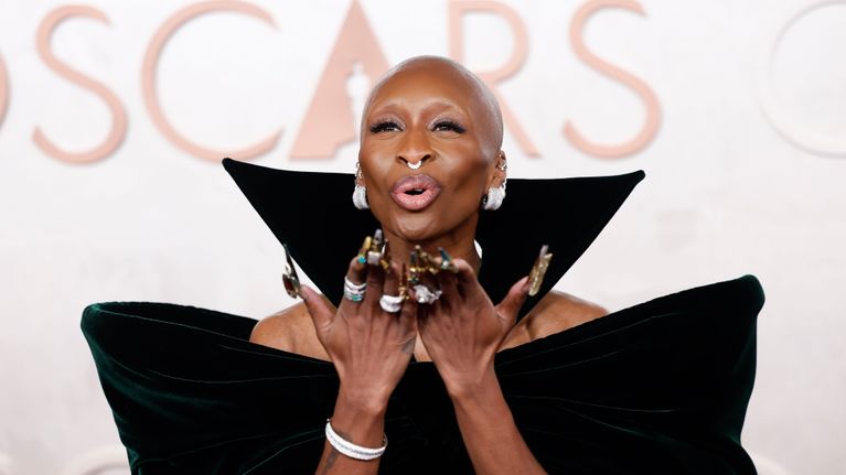 Cynthia Erivo puhaltaa lentosuukon. Erivo on kalju, näyttävät korvakorut, sormukset ja kynnet. Yllään tumman vihreä samettinen iltapuku, jossa pystyssä oleva kaulus.