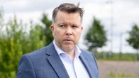Mies seisoo ulkona pukeutuneena siniseen pukuun ja vaaleaan paitaan, taustalla vihreitä puita ja harmaa taivas.